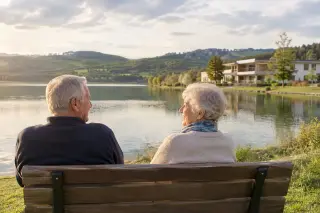 Deux seniors assis sur un banc face au lac de Pareloup avec une maison de retraite en arrière-plan et un paysage verdoyant sous une lumière douce.